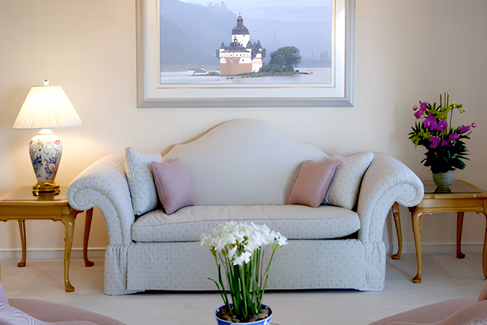 A cozy living room area with a light-colored sofa adorned with two pink pillows and two matching cushions. On either side of the sofa are wooden side tables, one with a decorative lamp and the other with a vase of purple flowers. A framed picture of a castle on a lake hangs on the wall above the sofa. In the foreground, there is a small pot of white flowers.