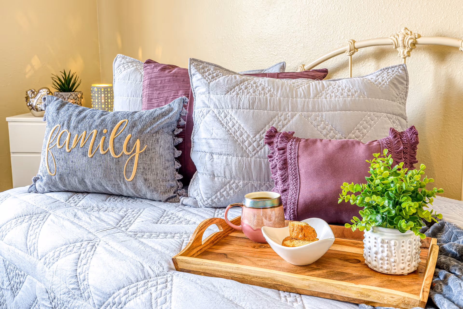 A cozy bed with a white quilted bedspread and multiple pillows, including a gray pillow with the word 'family' embroidered on it and purple pillows. On the bed is a wooden tray holding a ceramic mug, a white bowl with biscotti, and a small potted green plant. A white nightstand with a small plant and a decorative lamp is visible in the background.