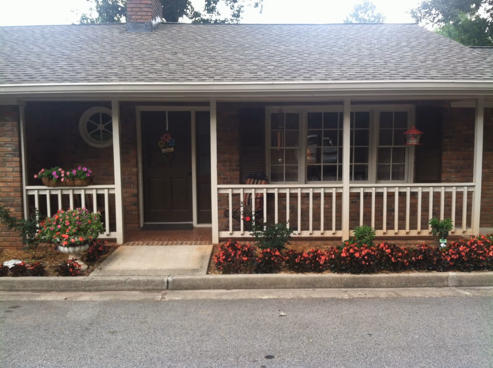 Brick building with a covered front porch, railing, windows, and flower beds.