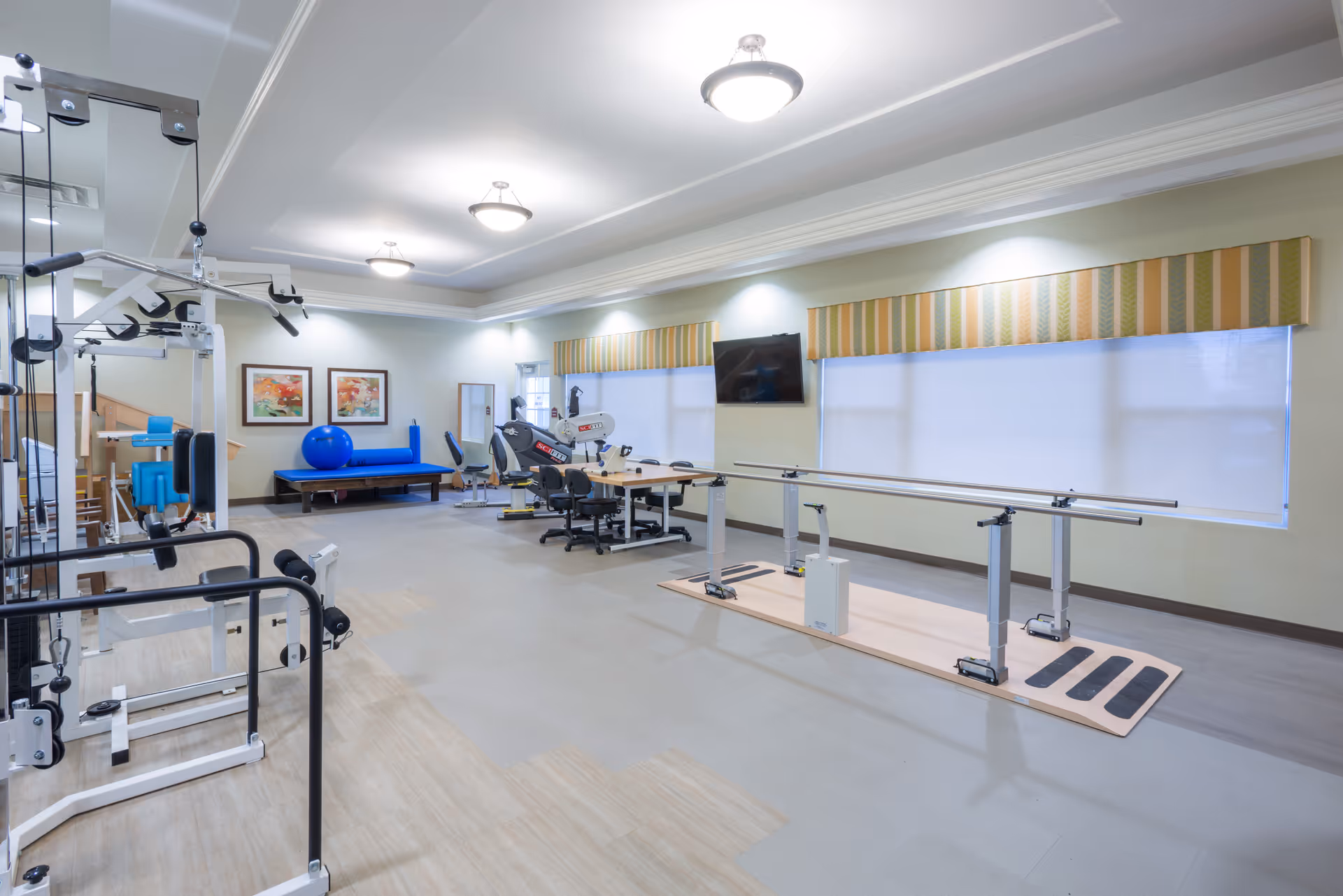 Senior living fitness room with parallel bars, exercise machines, and large windows.