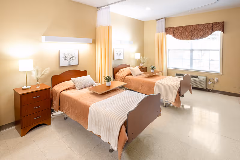 Bright shared patient bedroom with two hospital-style beds, bedside tables, lamps, and a large window.
