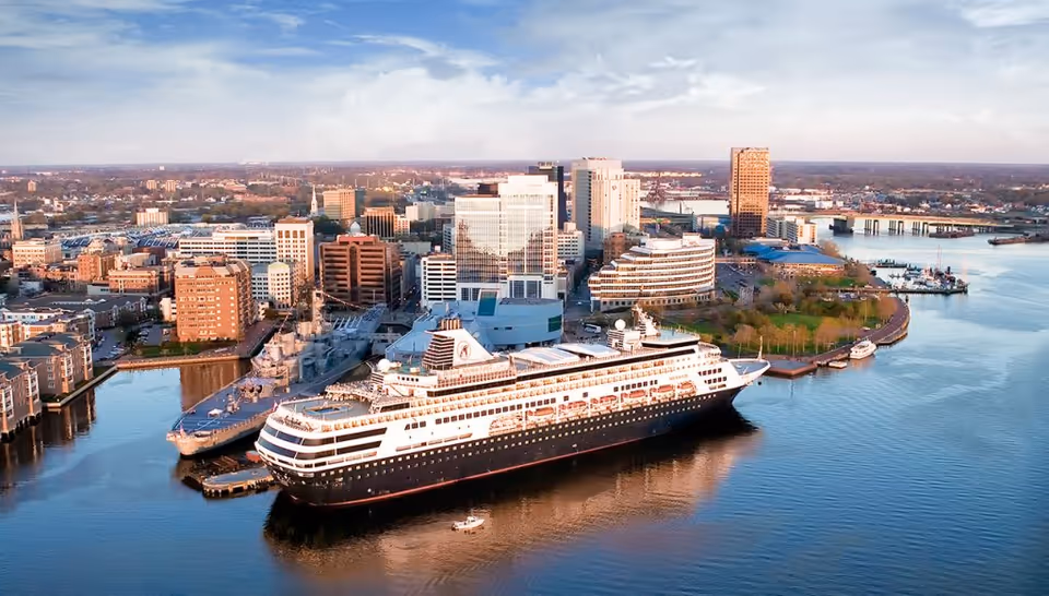 Aerial view of a waterfront cityscape featuring a large cruise ship docked at a pier, surrounded by various buildings and a calm body of water under a partly cloudy sky.