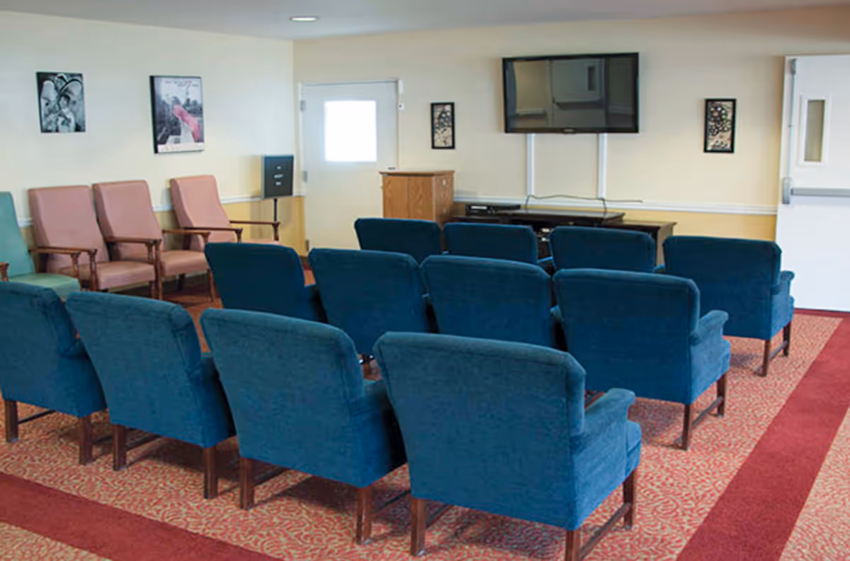 A small common area with rows of blue upholstered chairs facing a wall-mounted flat screen TV. There are additional pink and green chairs along the left wall, framed pictures on the walls, and a wooden cabinet beneath the TV. The room has a patterned carpet with red borders and light-colored walls.