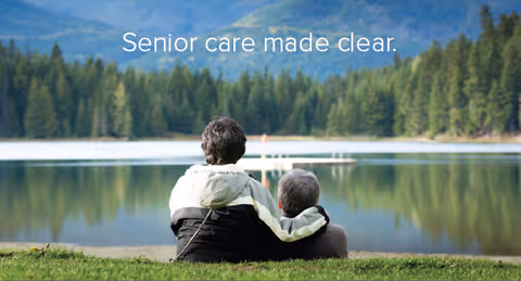 Two people sit on the grass by a calm lake with forested mountains behind.