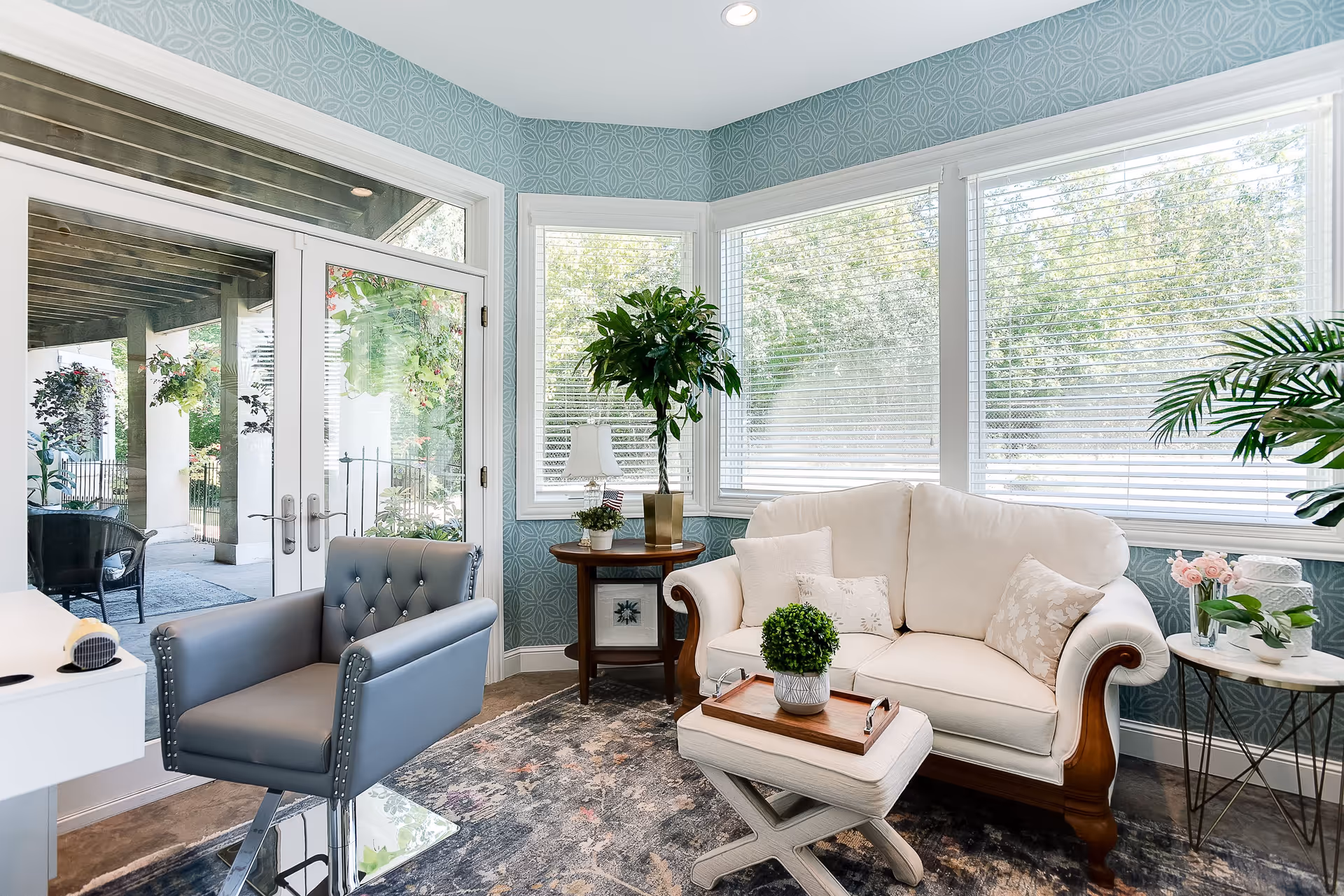 A bright and cozy living room area with large windows covered by white blinds, a cream-colored loveseat with decorative pillows, a small cushioned ottoman with a wooden tray and a potted plant on top, a blue armchair with studded detailing, a round wooden side table with a lamp and a tall potted plant, and a small round table with flowers and another plant. Outside the glass doors, there is a covered patio with hanging plants and outdoor seating.