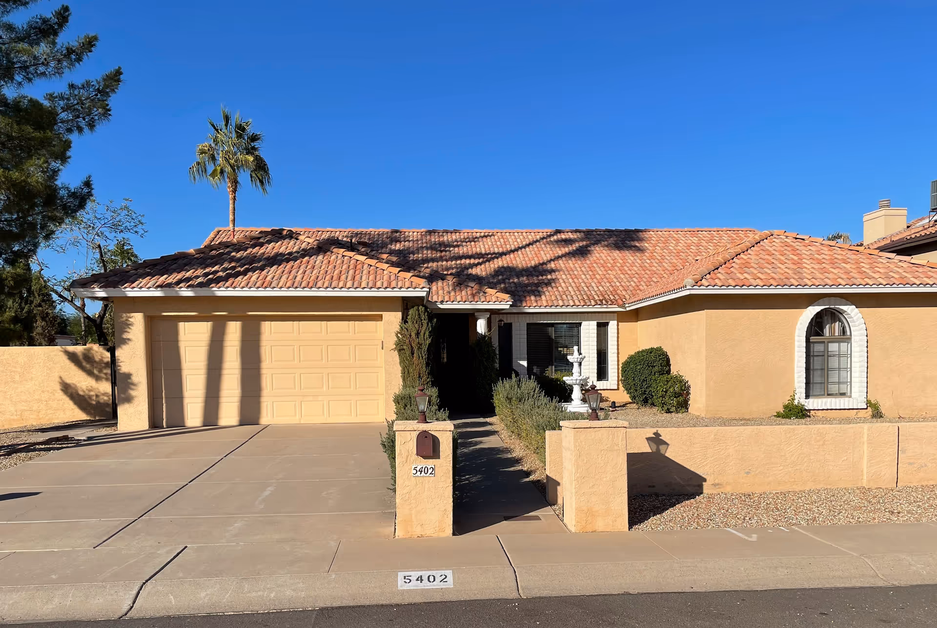 Single-story stucco house with a tiled roof, two-car garage, driveway and mailbox numbered 5402 in a sunny front yard.