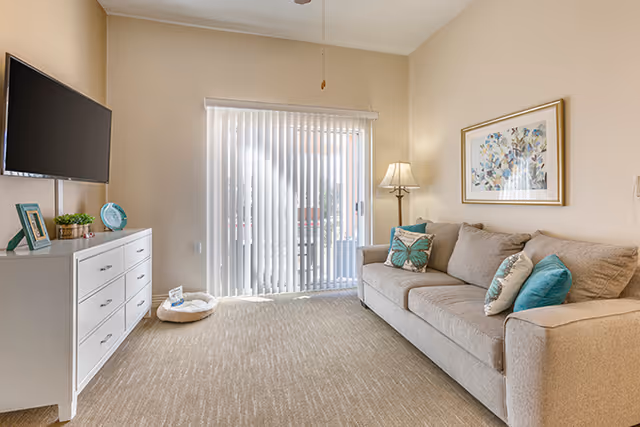 Cozy living room in a senior living facility with sofa, TV, dresser, and sliding glass doors.