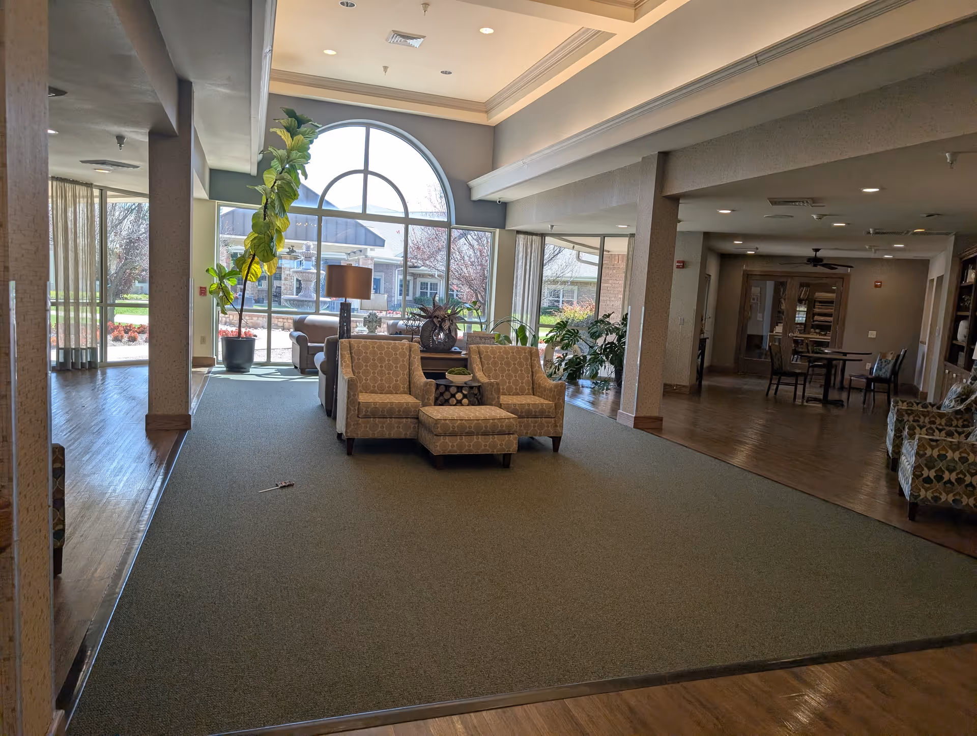 Bright senior living lounge with arched window, comfy chairs, plants, and wooden flooring.