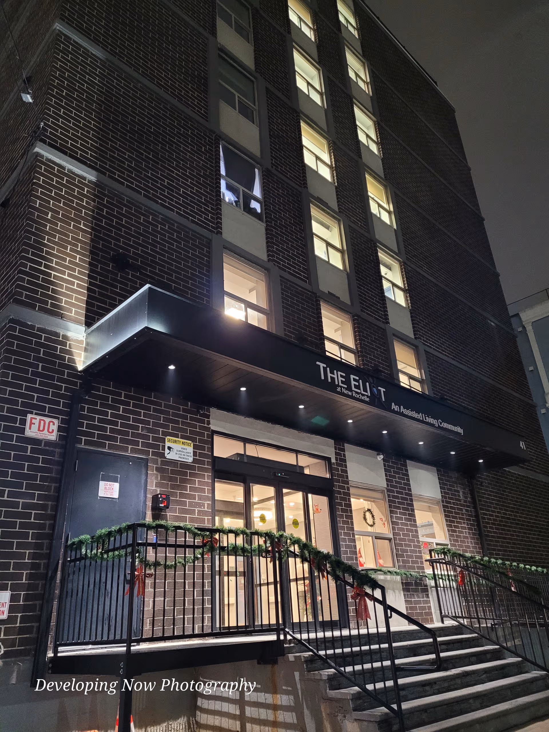 Nighttime exterior of a brick senior living building with decorated entrance.
