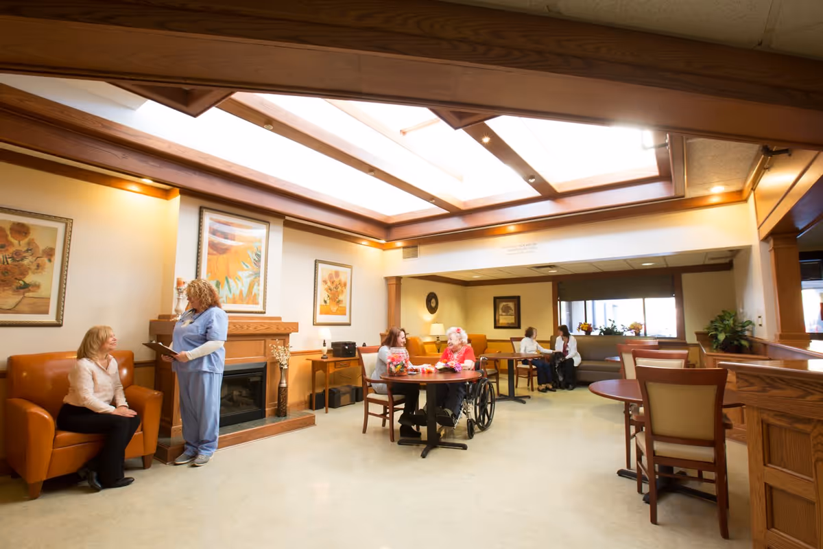 A bright and spacious common area in a senior living facility with a large skylight ceiling. Several elderly residents and caregivers are seated and engaged in conversation around tables and chairs. The room features warm wood accents, framed floral artwork on the walls, and a fireplace with a wooden mantel.
