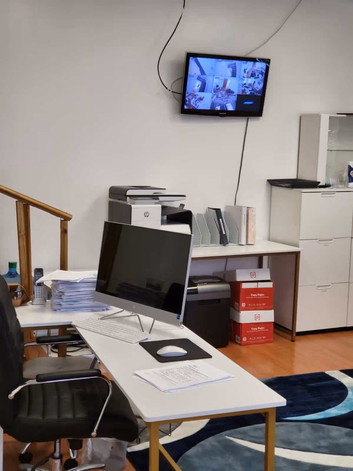 A tidy office desk with computer, mouse, paperwork, and a wall-mounted monitor displaying security feeds.
