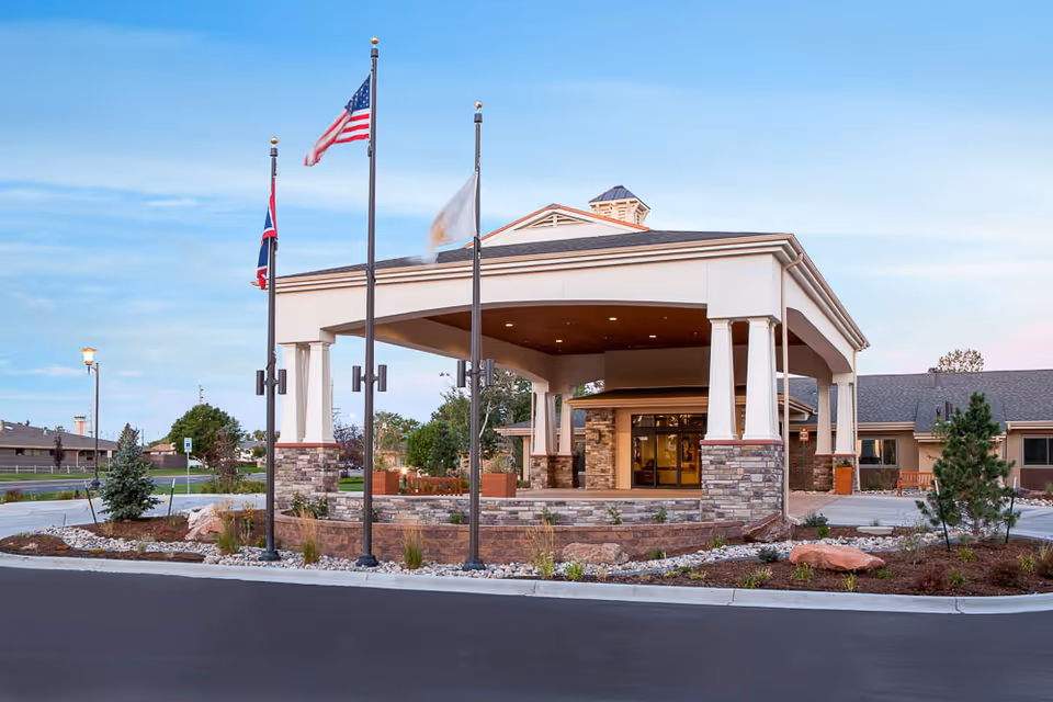 Front entrance of Life Care Center of Cheyenne featuring a covered drop-off area with stone pillars, three flagpoles with flags, landscaped garden beds, and a clear sky background.