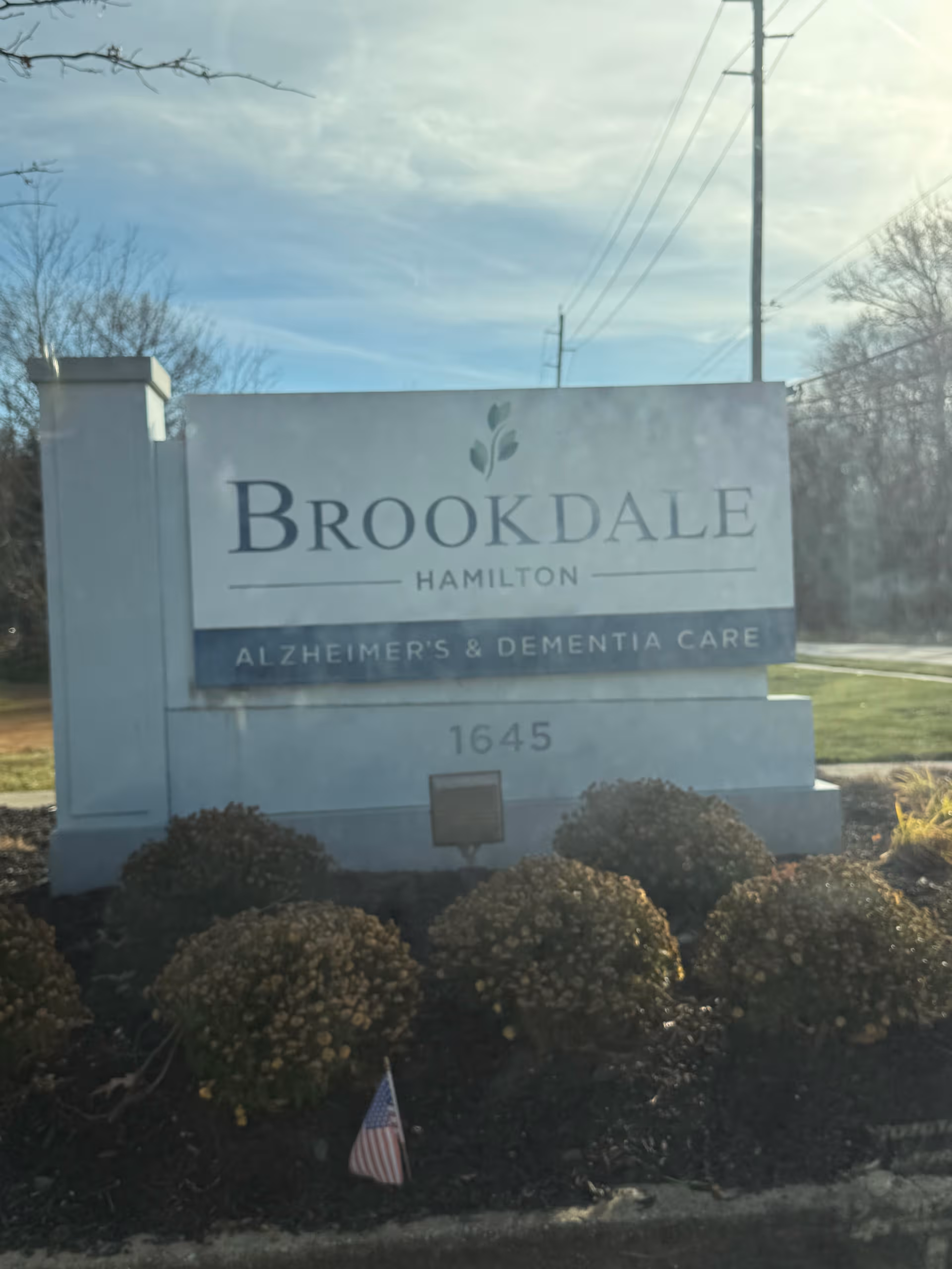 Brookdale Hamilton sign at entrance with shrubs and a small American flag