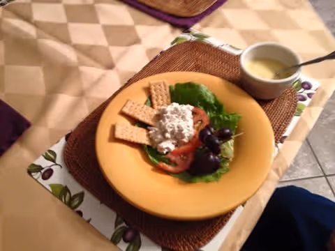 A yellow plate with a salad consisting of lettuce, tomato slices, grapes, a scoop of cottage cheese, and several rectangular crackers on a woven placemat. Next to the plate is a small white bowl with a spoon, containing a creamy dressing or sauce. The setting is on a table with a beige tablecloth and a decorative olive branch patterned runner.