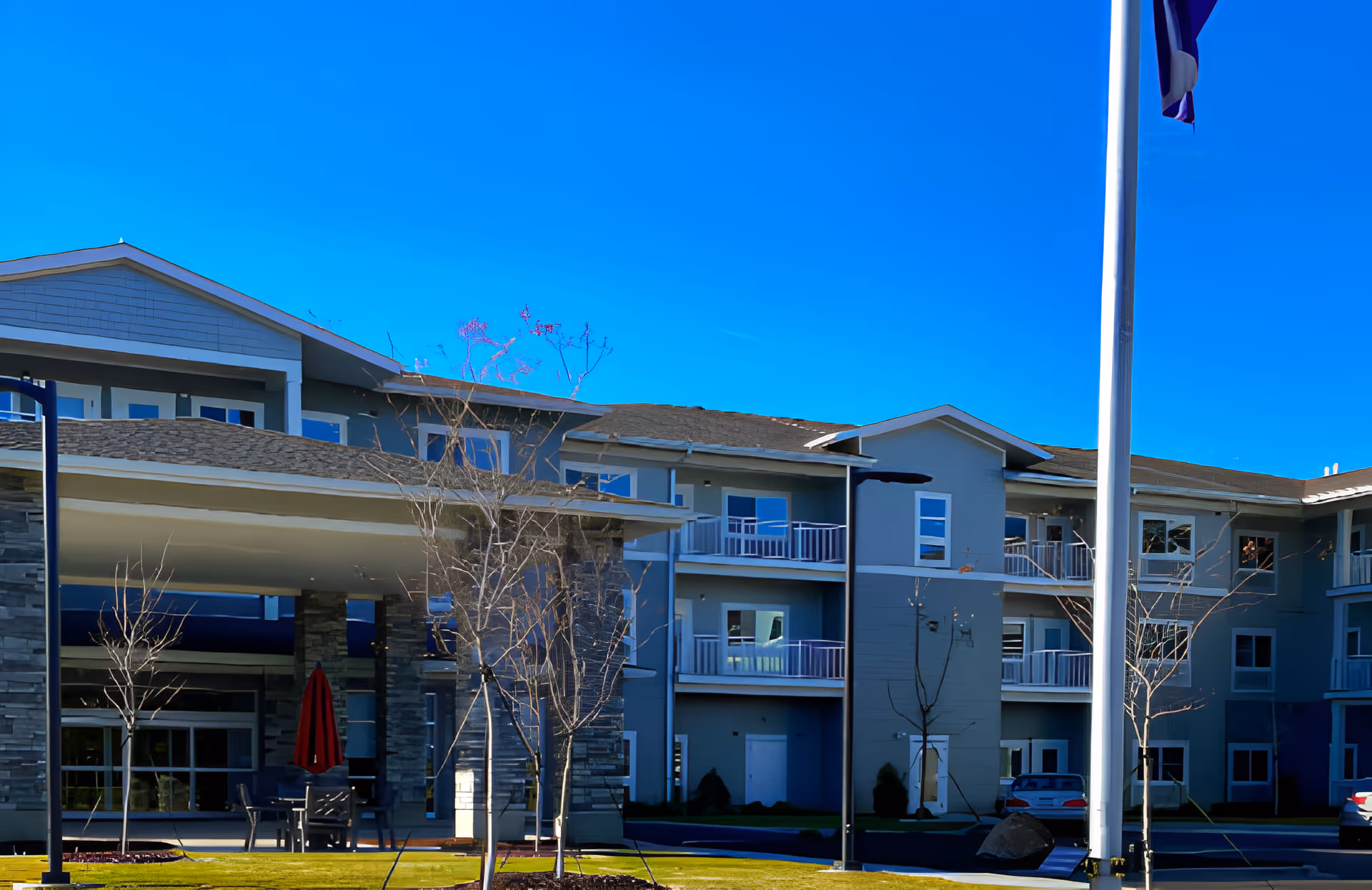Exterior view of a multi-story retirement community building with balconies, a covered entrance supported by stone pillars, a flagpole, and a clear blue sky.