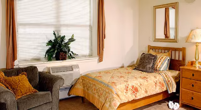 Cozy bedroom in a senior facility featuring a bed, sofa, dresser, window, and plant.