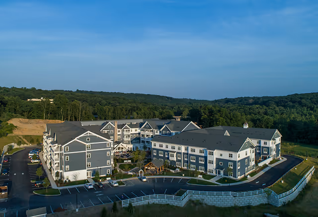 Aerial view of a large senior living facility building with multiple wings, surrounded by parking lots and greenery, under a clear blue sky.