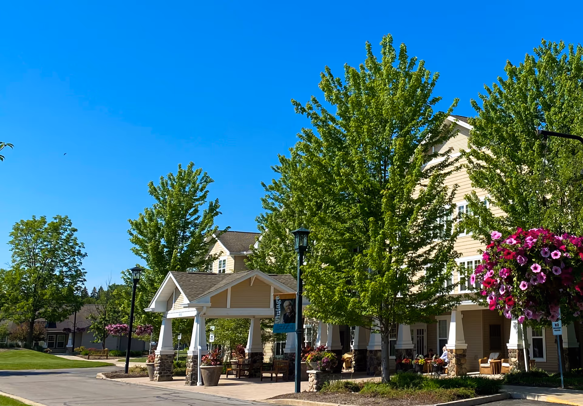 Front entrance of a senior living building with a covered porte-cochere, large trees, and hanging flower baskets under a clear blue sky.