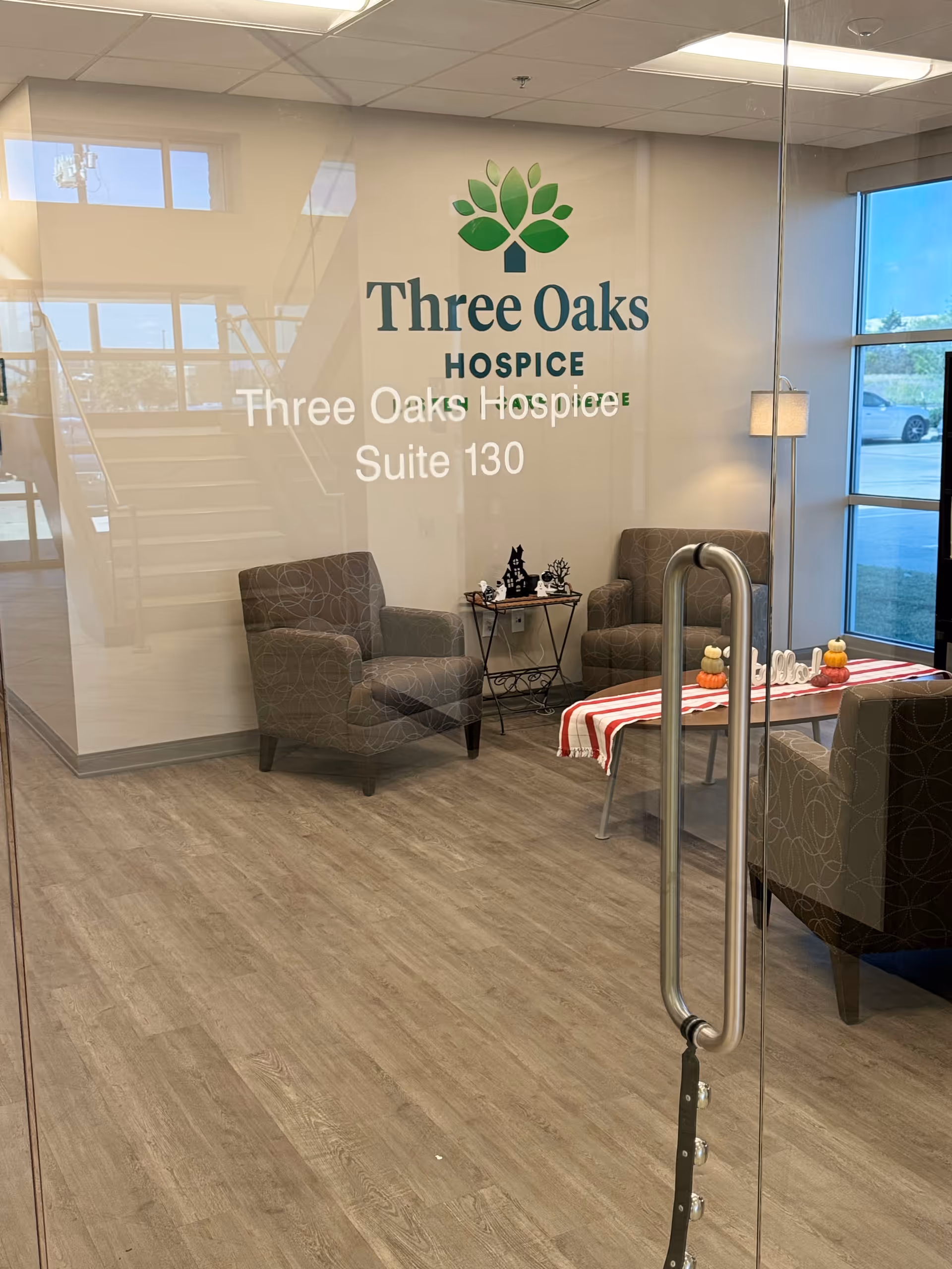 Glass-walled lobby with Three Oaks Hospice logo, seating, and a small table.