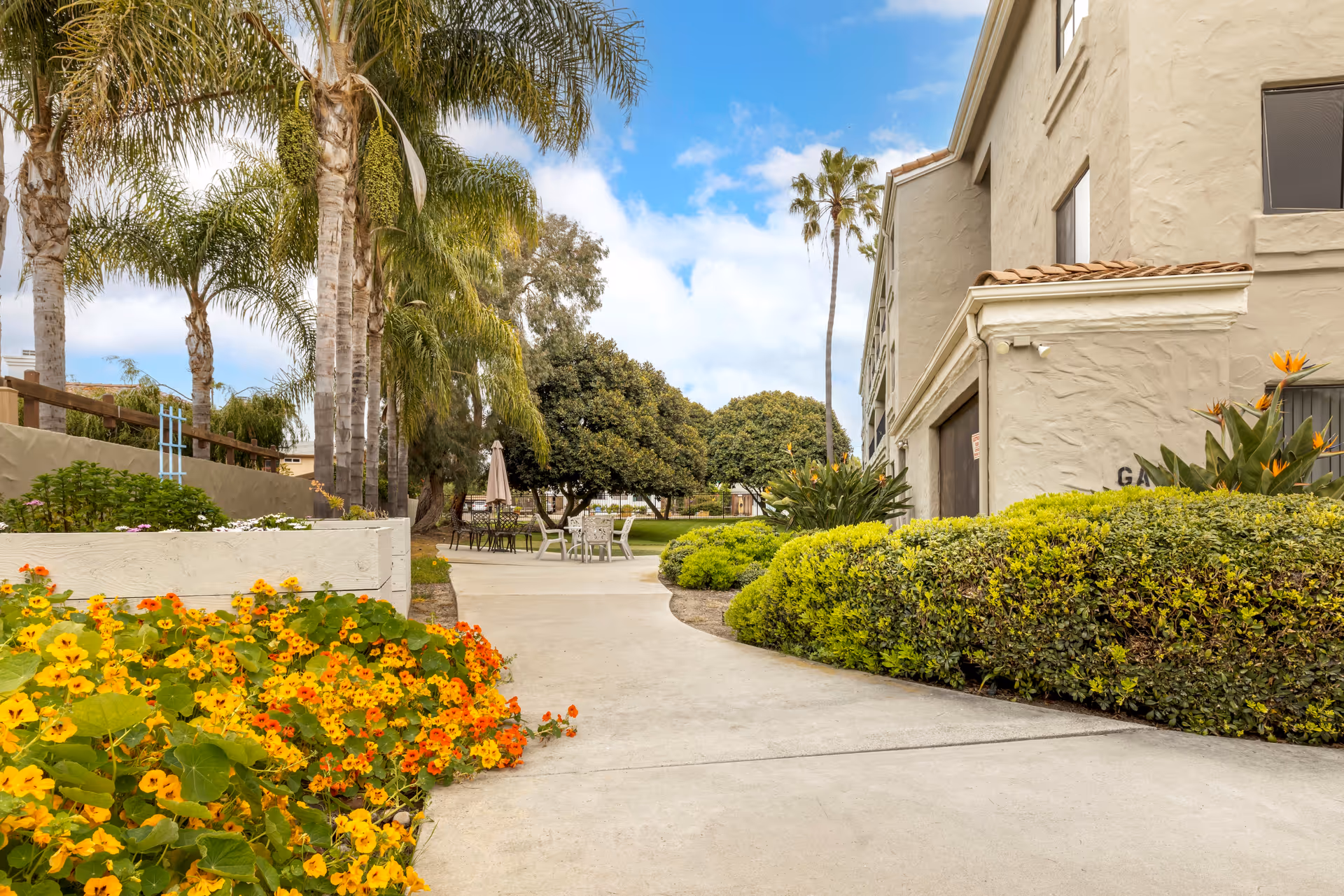 Sunny outdoor courtyard with palm trees, flowers, and seating beside a beige senior living building.