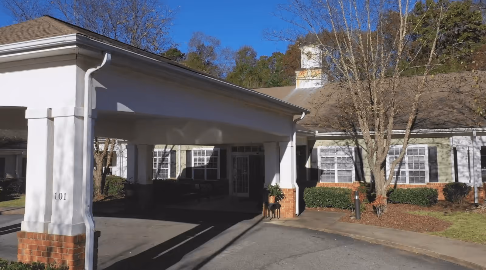 Exterior view of a senior living facility building with a covered entrance supported by white columns with brick bases. The building has multiple windows with white shutters, a tree without leaves, and some landscaping with bushes and mulch. The sky is clear and blue.