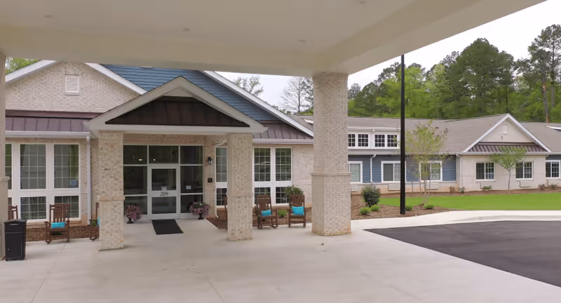 Entrance of The Colonial at Old Camden senior living facility showing a covered drop-off area with brick pillars, a paved driveway, and a building facade with large windows and a peaked roof. There are chairs with blue cushions and some potted plants near the entrance.