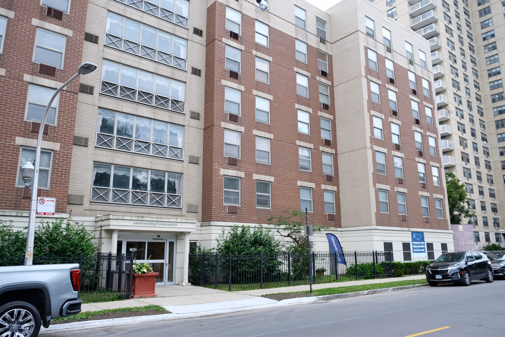 Front exterior of a multi-story brick senior living building with an entrance, sidewalk, parked cars, and landscaping.