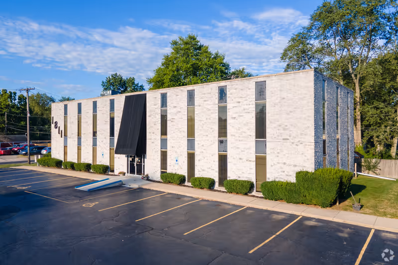 Two-story brick senior living building with tall windows, black entrance awning, and a parking lot.