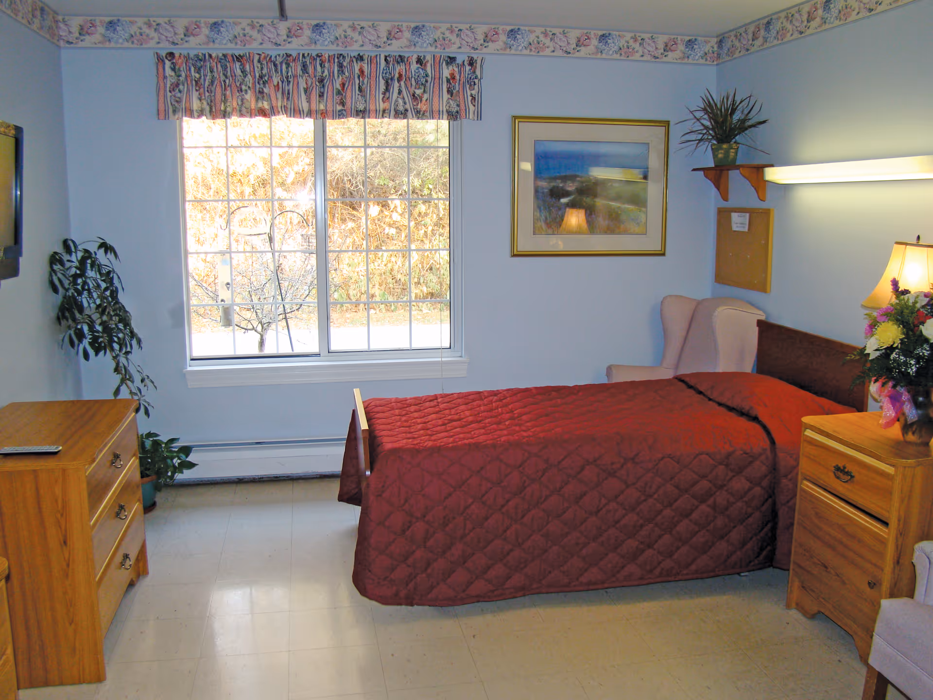 A simple bedroom in a senior living facility with a single bed covered in a red quilt. The room has light blue walls with floral wallpaper border near the ceiling. There is a wooden dresser with a remote control on top, a potted plant on the floor, a window with floral valance curtains, a framed picture on the wall, a pink armchair, a wooden nightstand with a lamp and a flower arrangement, and a small shelf with a potted plant above the armchair.