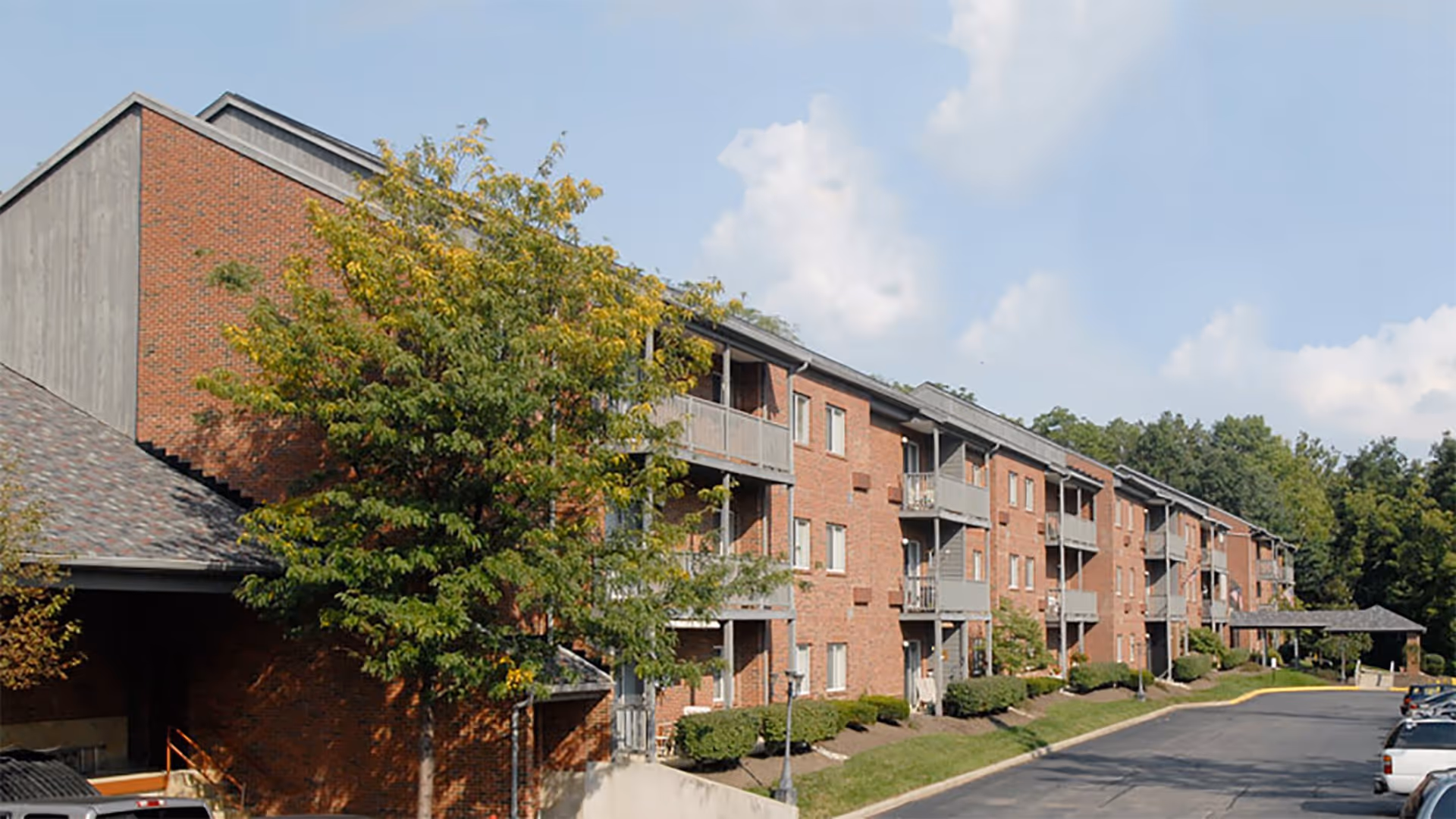 Exterior front view of a multi-story red brick retirement apartment building with balconies, trees, and a driveway.