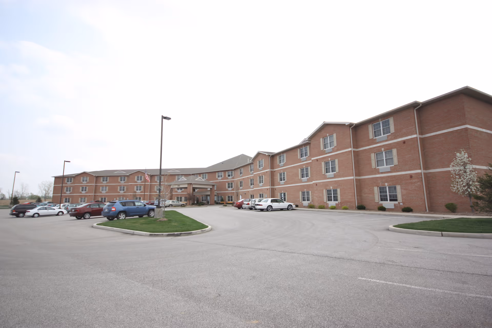 Wide-angle view of a three-story brick senior living building with a circular driveway and parked cars.