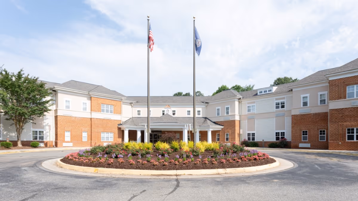 Front exterior view of Morningside of Bellgrade, a two-story senior living facility with a circular driveway and landscaped flower bed in the center featuring two flagpoles with the American flag and another flag.
