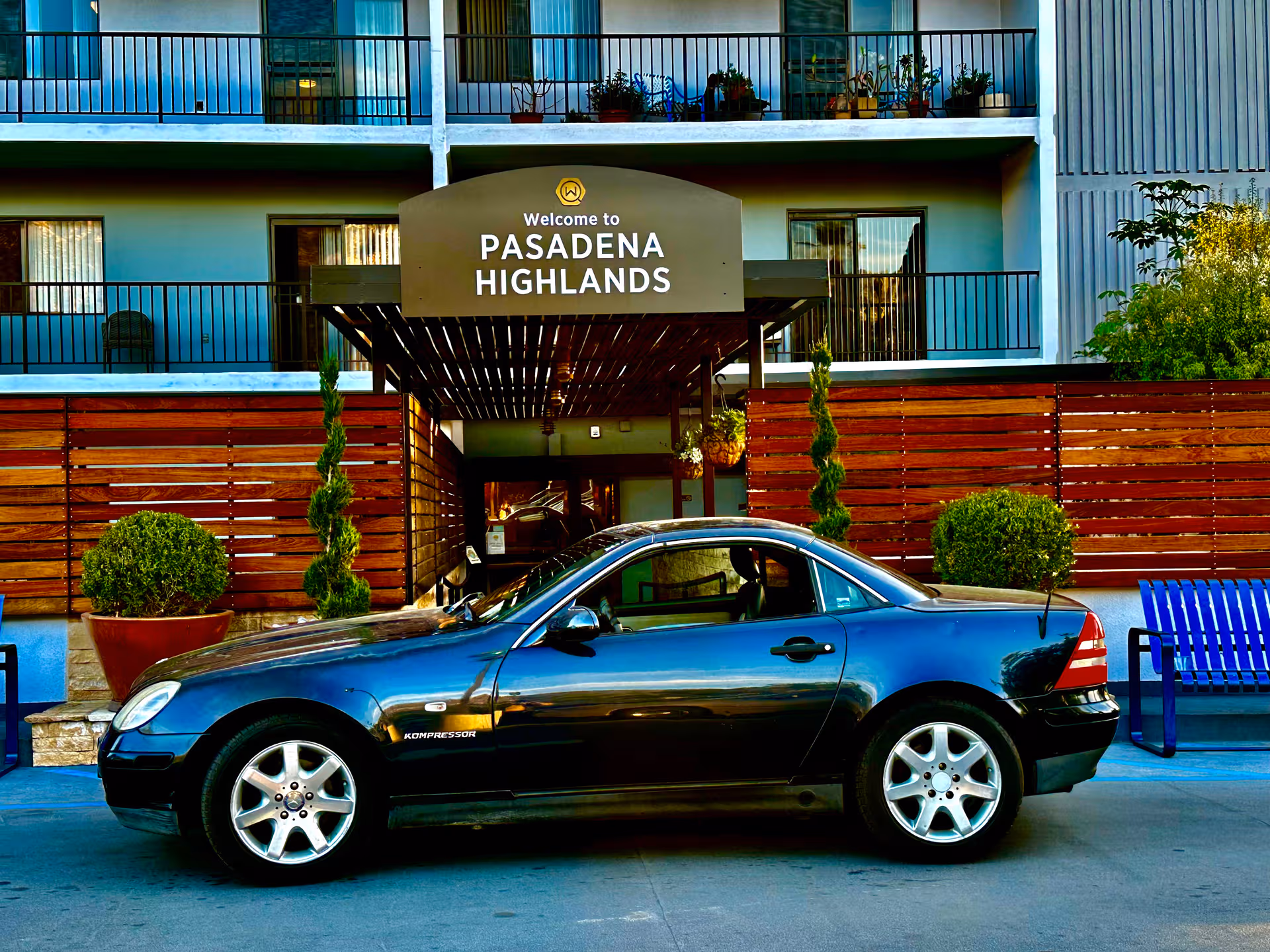 Front of Pasadena Highlands with sign, wooden fence, and a blue car parked outside.