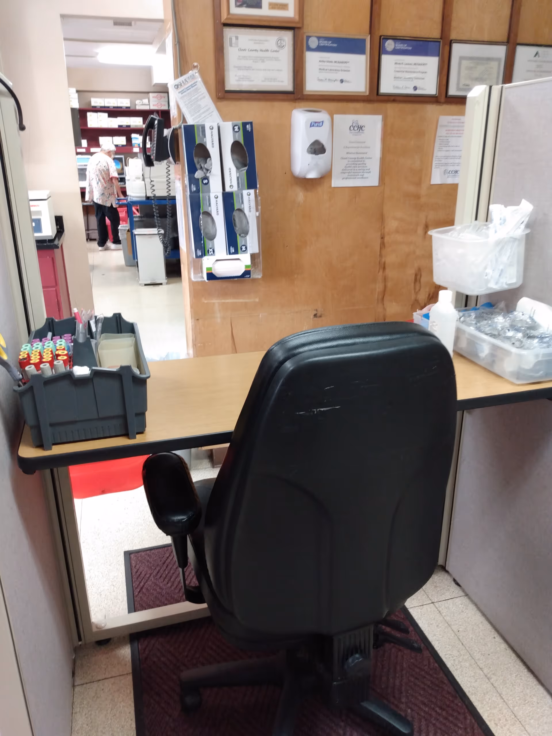 Reception desk with chair, supplies, certificates on wall, and staff in background