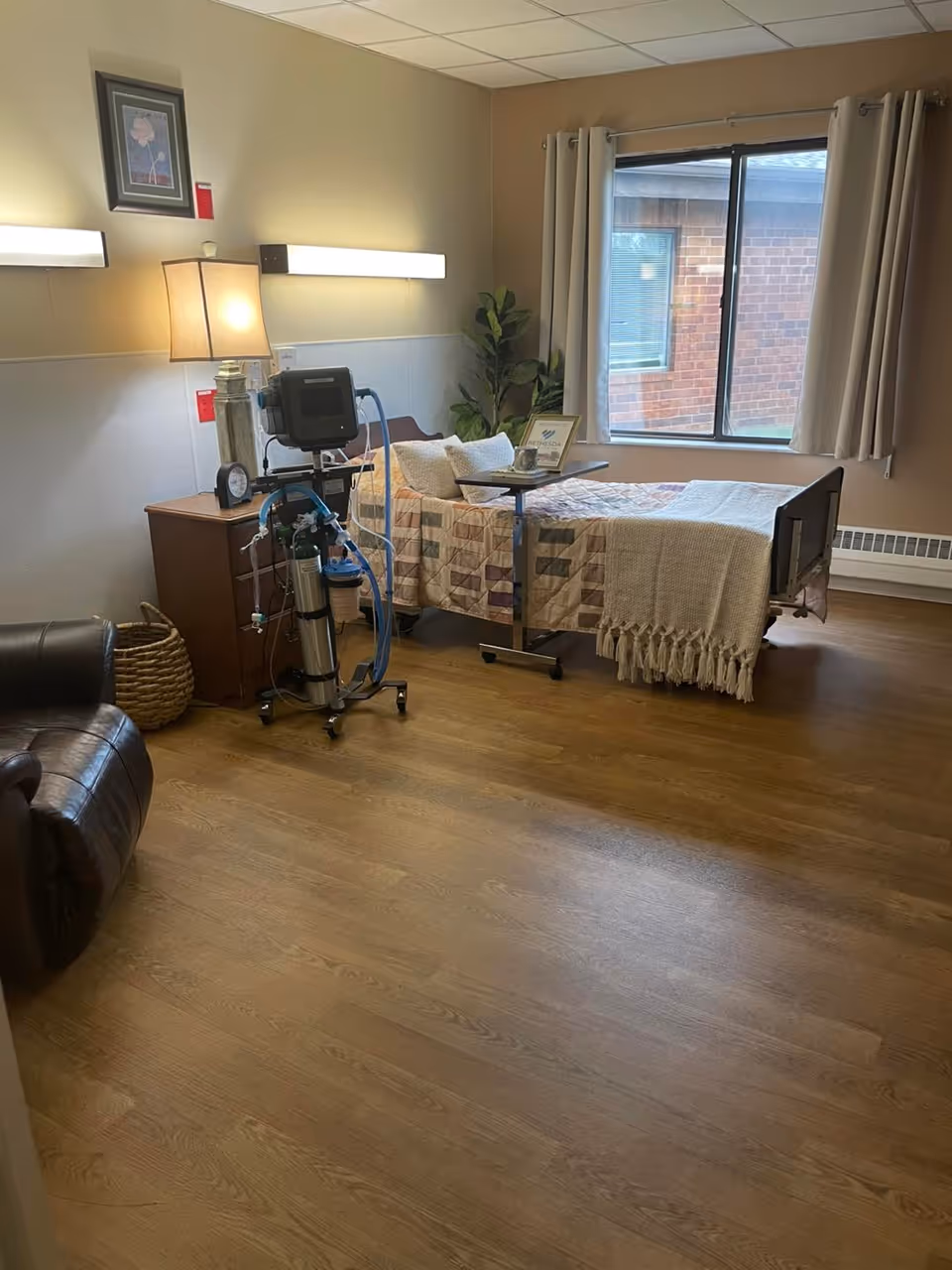 A cozy senior care bedroom with a hospital-style bed covered with a patchwork quilt and a beige throw blanket. Next to the bed is a wooden nightstand with a lamp and medical equipment, including an oxygen tank and monitor. A small overbed table holds a cup and a framed picture. The room has a large window with beige curtains, a potted plant in the corner, and a comfortable leather recliner chair. The floor is wooden, and the walls are painted in warm neutral tones.