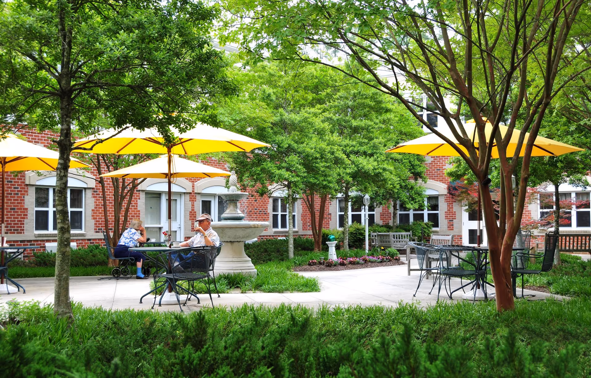 Outdoor courtyard area at The Crossings at Ironbridge with green trees, yellow umbrellas shading metal tables and chairs, a stone fountain, and two elderly people sitting and conversing.