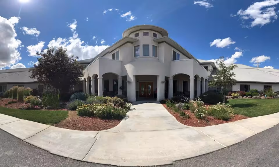 Front entrance of a senior living building with an arched portico, landscaped flower beds, and a bright blue sky.
