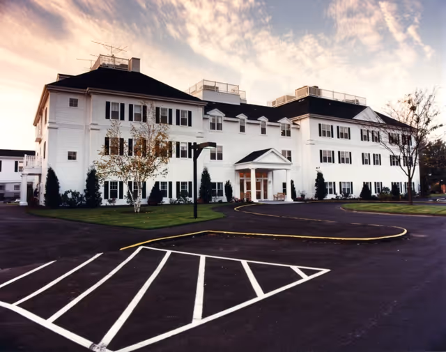Front exterior of a large white multi-story building with a circular driveway, parking spaces, and landscaped grounds under a cloudy sky.