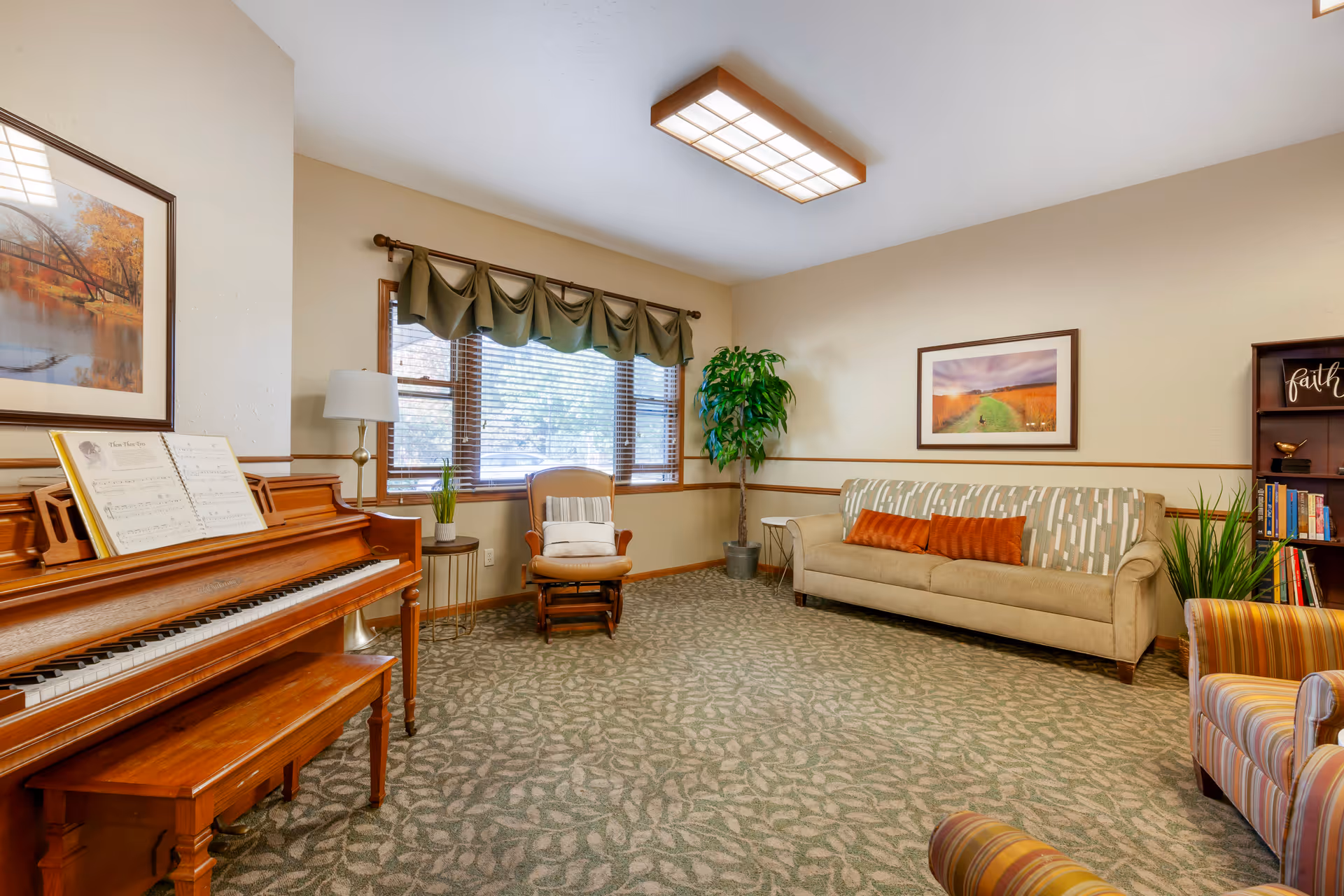 Sunny senior living lounge with piano, sofa, chairs, plants, and framed wall art.