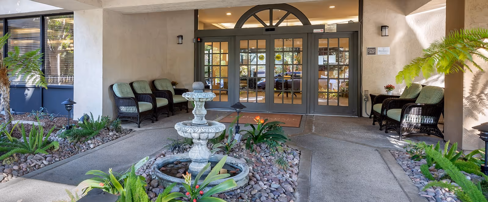 Entrance area of a senior living facility with a decorative stone fountain surrounded by plants and rocks in the center. There are cushioned wicker chairs arranged along the walls on both sides of the entrance. The entrance features double glass doors with a semi-circular window above them, allowing a view into the interior.