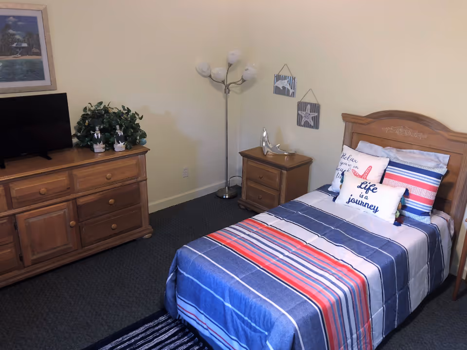 A single bedroom in a senior living unit with a striped bed, dresser, and TV.