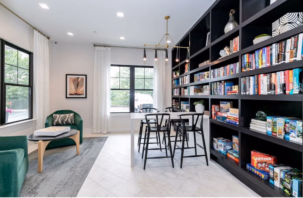 A bright and modern common area with large windows, white curtains, and a black bookshelf filled with books and board games. There is a white table with four black chairs in front of the bookshelf, and two green armchairs with a small round table between them on a gray rug.