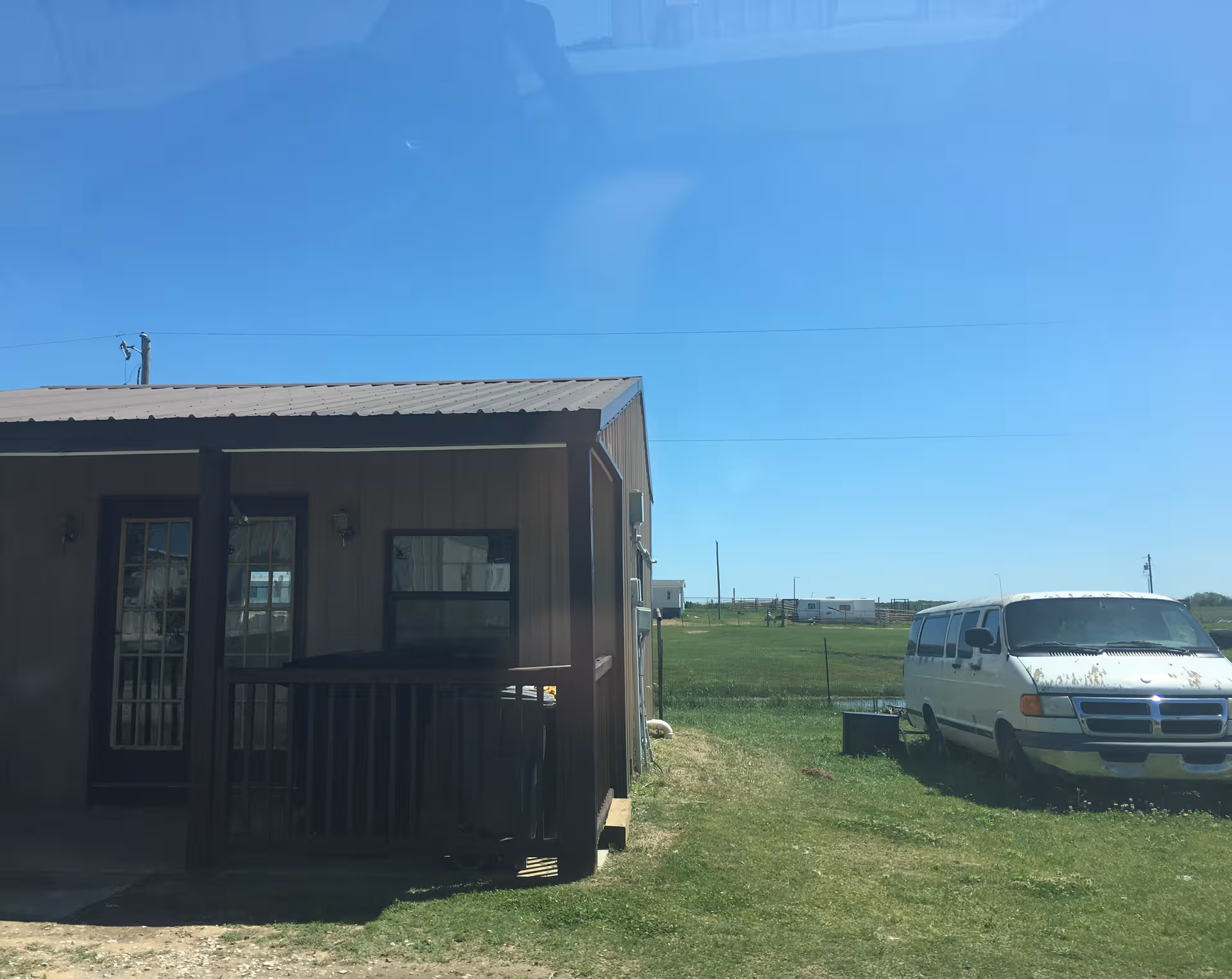 Small brown building with porch and railing beside a grassy field and white van.