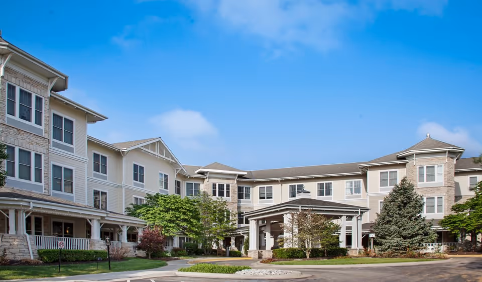Front exterior of a multi-story senior living building with a covered entrance, landscaping, and a clear blue sky.