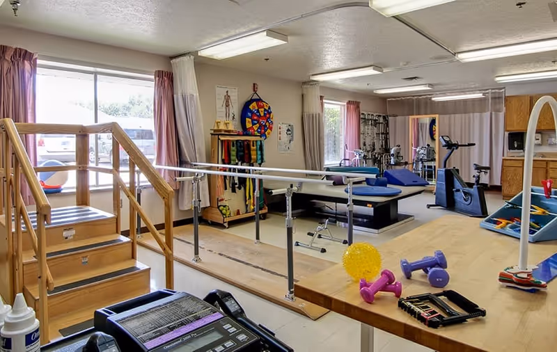 Physical therapy room with wooden stairs and handrails for rehabilitation exercises, parallel bars, exercise equipment including stationary bike, therapy tables, resistance bands, weights, and therapy balls. Large windows with curtains provide natural light.