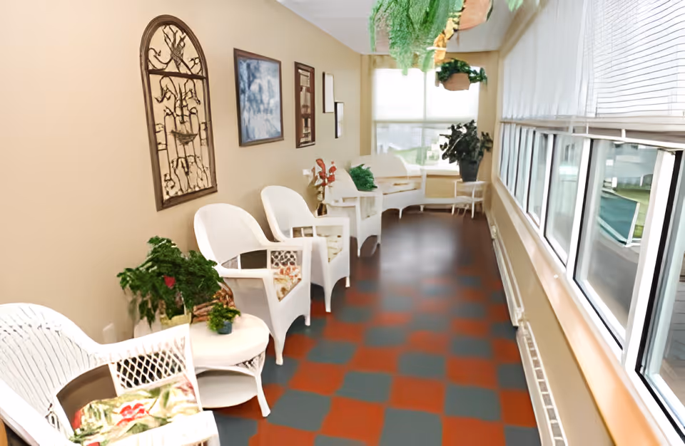 A bright indoor sitting area with white wicker chairs and small tables arranged along a beige wall adorned with framed artwork and decorative wall hangings. The floor has a checkered pattern in red and gray, and large windows with blinds line the opposite wall, letting in natural light. Several potted plants are placed on tables and hanging from the ceiling, adding greenery to the space.