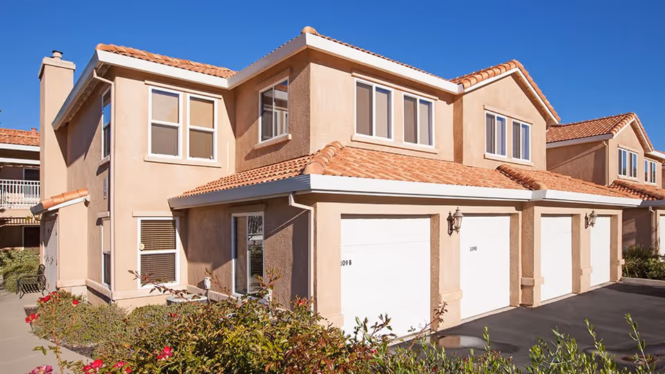 Exterior view of a two-story residential building with beige stucco walls and red tile roofing. The building features multiple windows and three white garage doors. There are some bushes with red flowers in front of the building and a clear blue sky overhead.