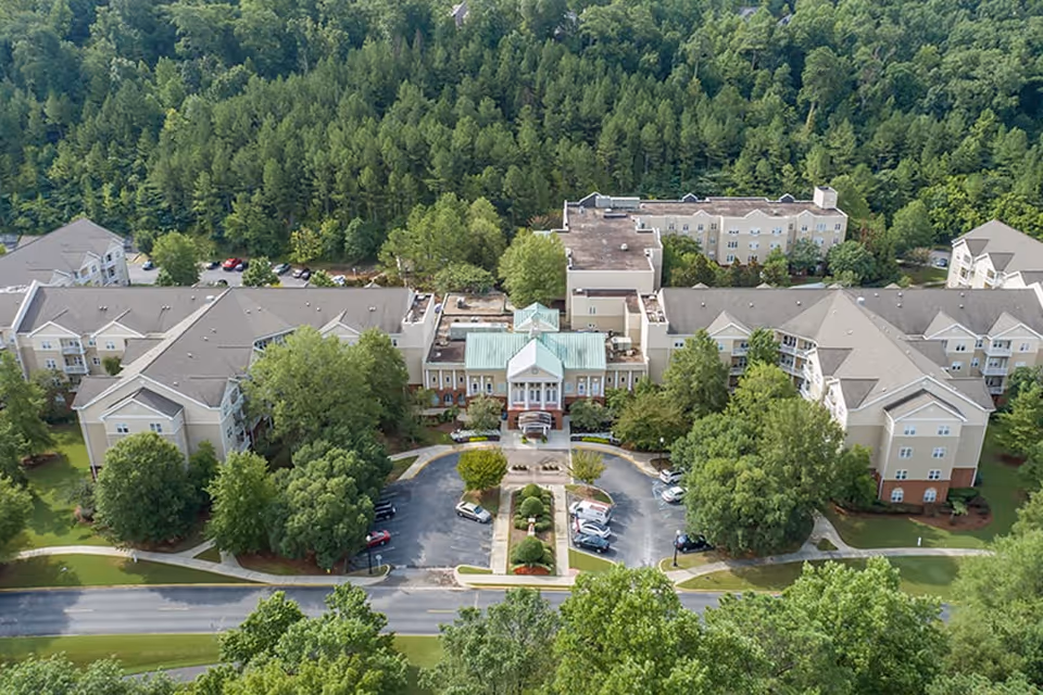 Aerial view of Brookdale University Park senior living facility surrounded by dense green trees. The building complex has multiple connected beige structures with gray roofs and a central entrance with a green roof. There is a parking lot with several cars in front of the entrance and a road in the foreground.