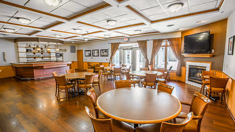 A spacious dining room with multiple round wooden tables and chairs arranged neatly. There is a bar area labeled 'PUB' on the left side with shelves holding glasses and bottles. The room has large windows with brown curtains allowing natural light to enter. A flat-screen TV is mounted above a fireplace on the right wall. The ceiling features recessed lighting and decorative panels.
