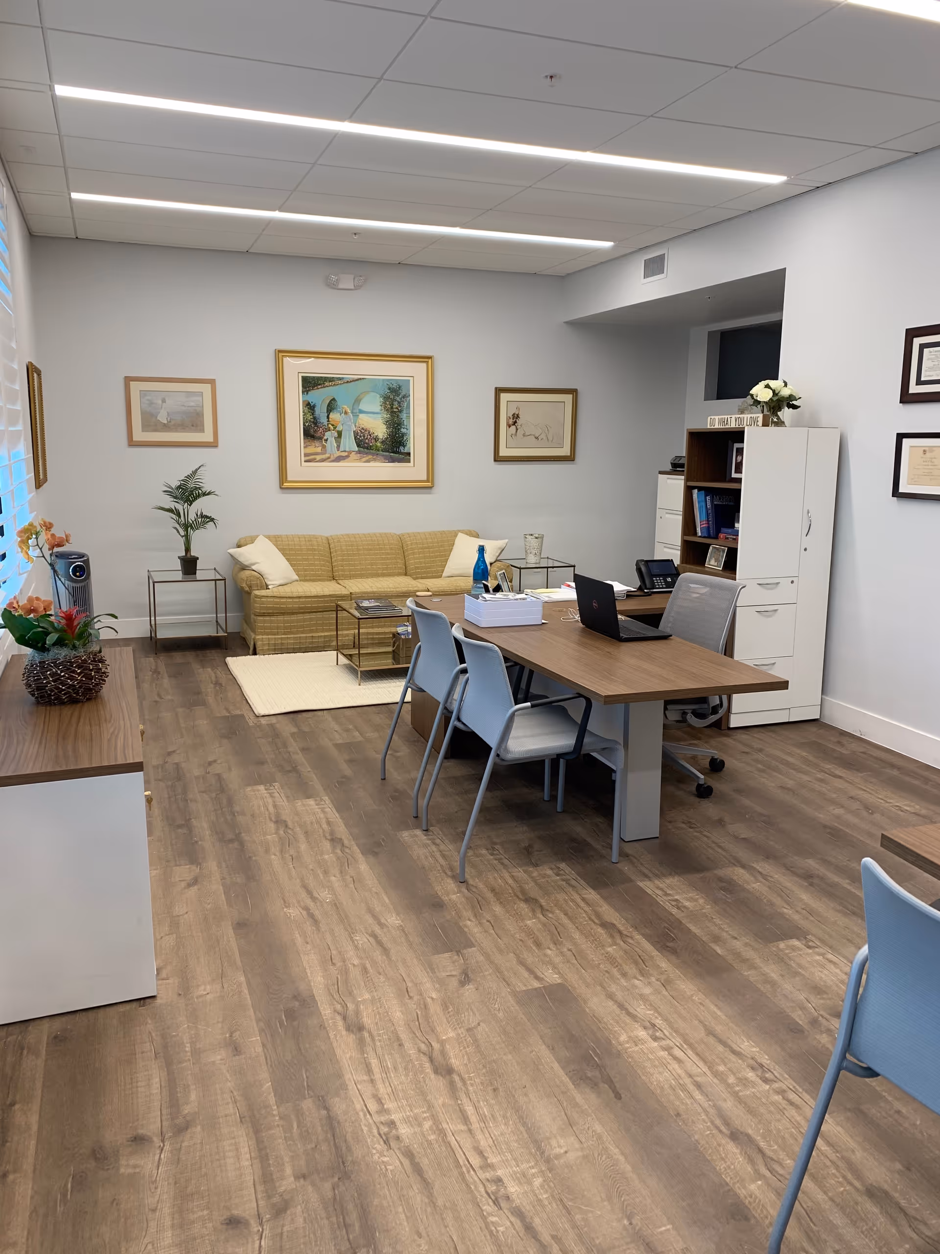Bright, softly furnished common area with a sofa, desk, chairs, bookshelf, and artwork.